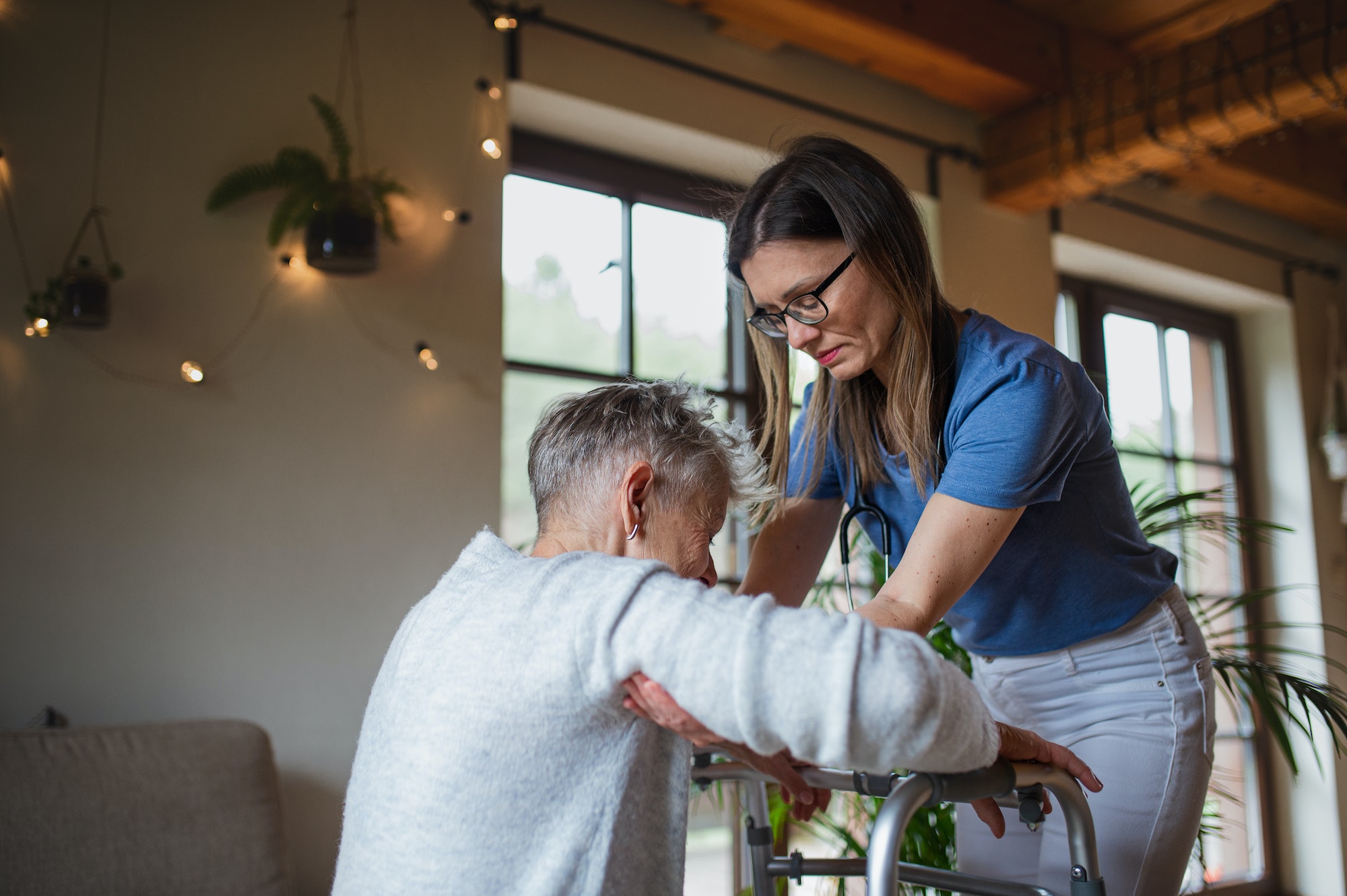 Healthcare worker or caregiver visiting senior woman indoors at home, helping her to walk